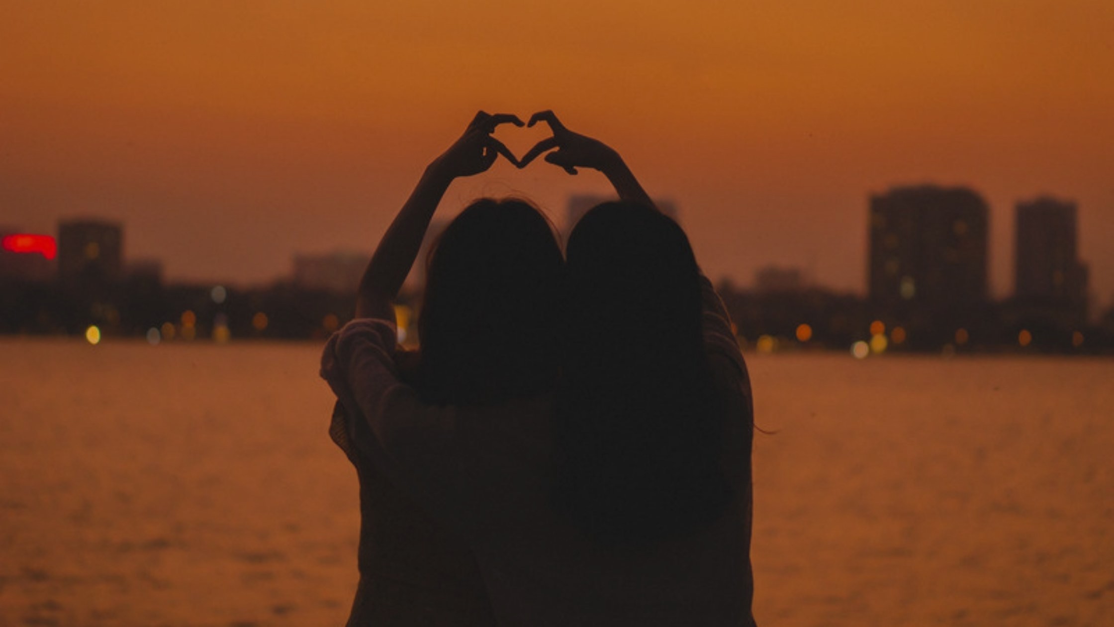 Two friends making a heart with their hands over the water at sunset