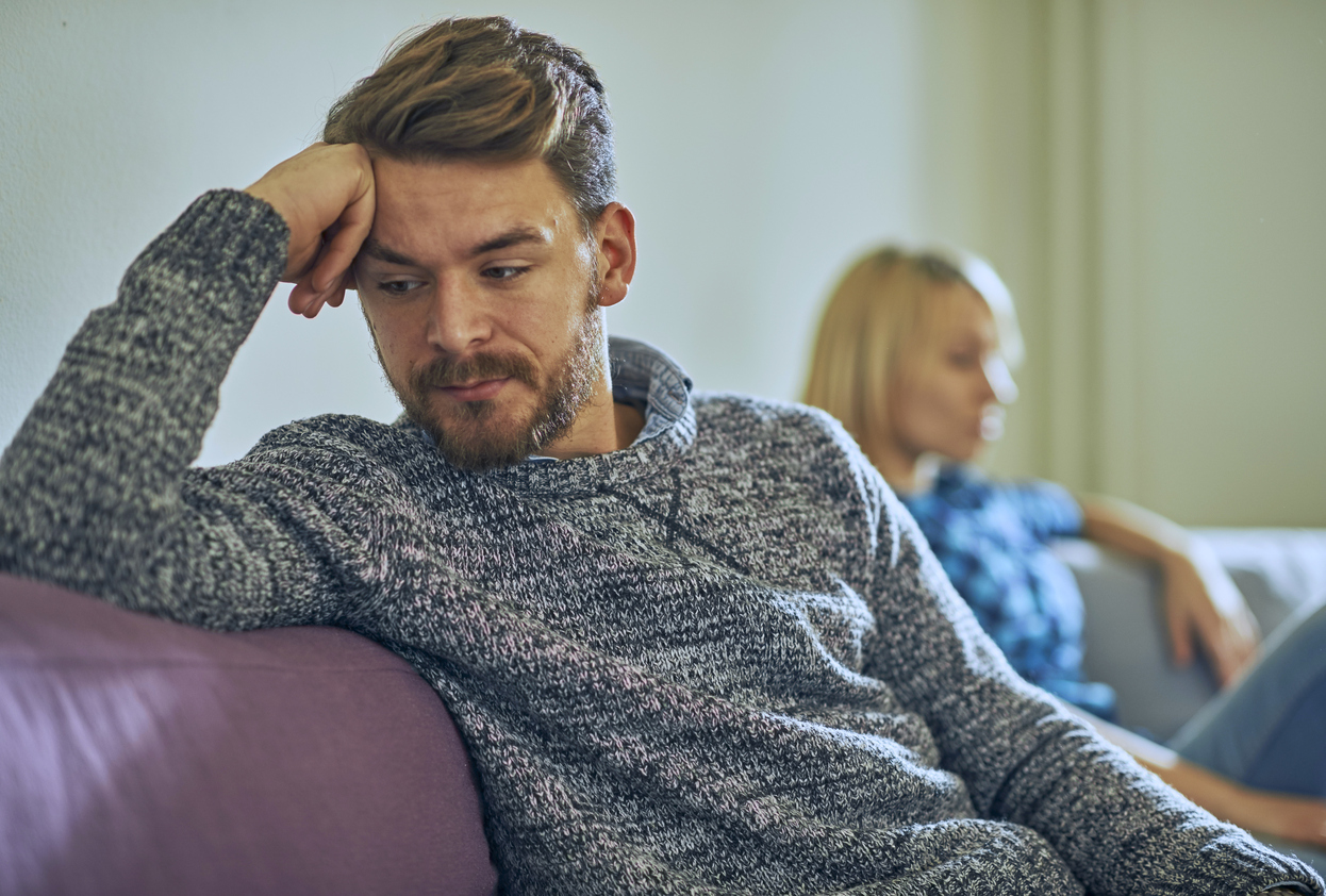 Man looking down and withdrawn on the couch while his partner sits behind him